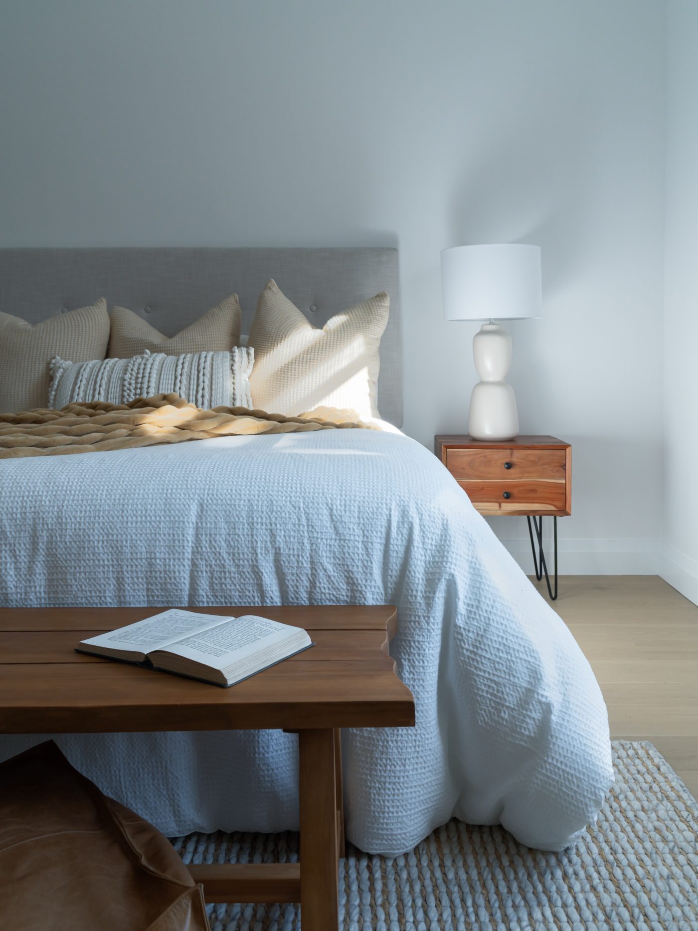 Bedroom in soft window light with white waffle-weave bedding, a tan throw, and a wood bench with an open book in the foreground