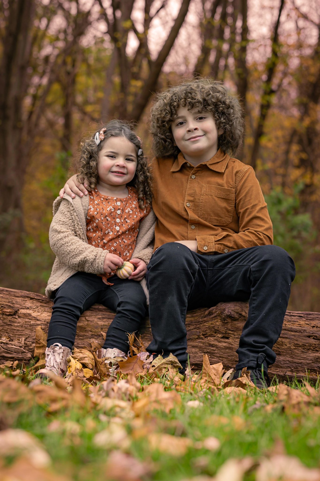 Two siblings sitting together on a fallen log in autumn woods, the older brother with his arm around his little sister, who holds a small pumpkin